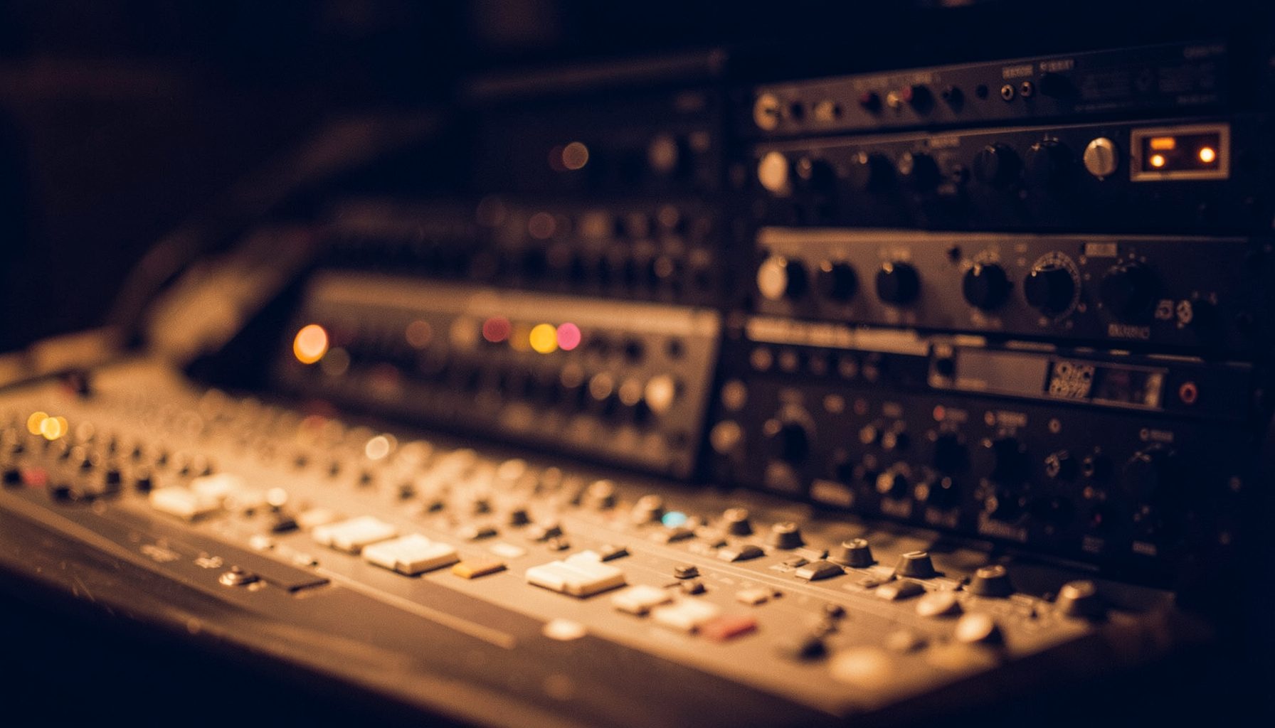 Warm amber studio lighting across a mixing console with rack-mounted outboard gear in soft focus, deep navy shadows