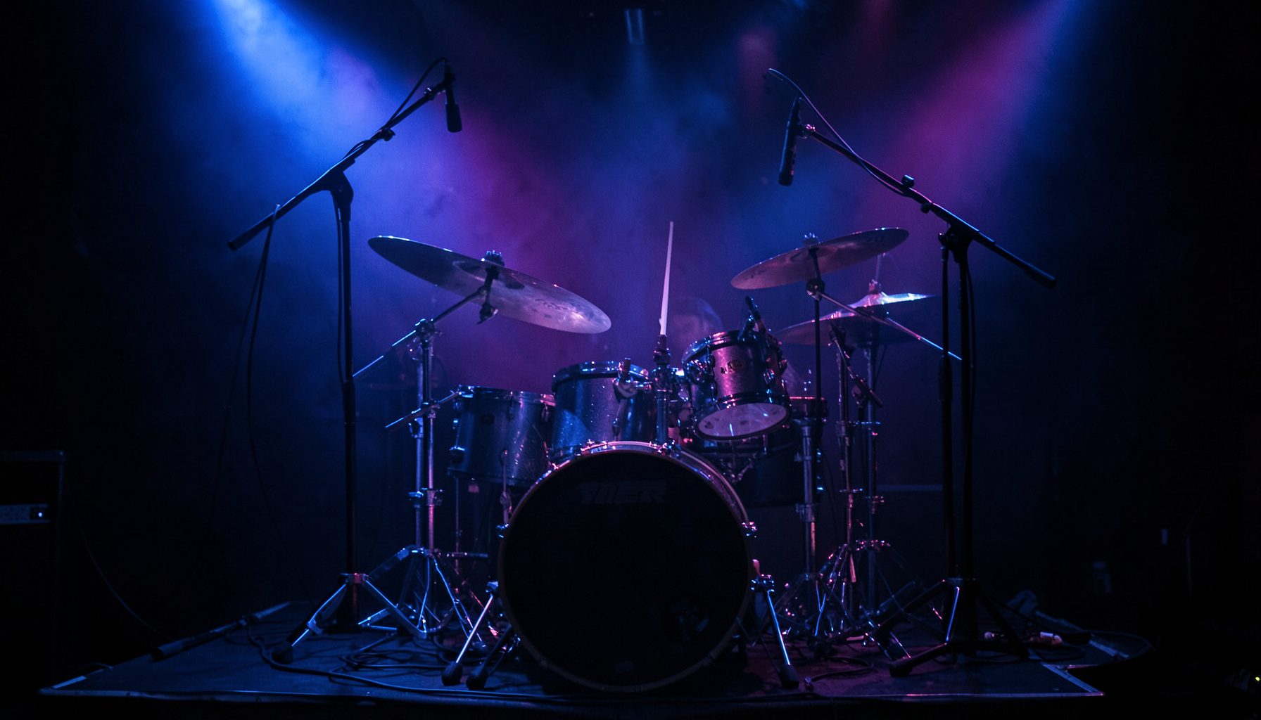 Dim stage lighting in deep blue and violet tones washing over a drum kit and microphone stands, moody and atmospheric