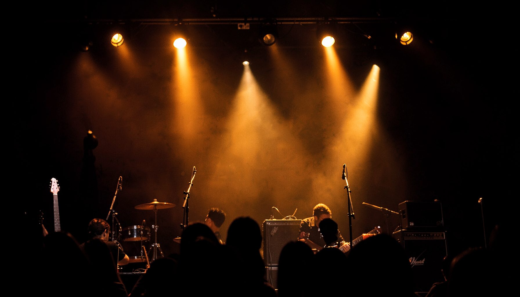 Warm amber and gold stage lights glowing against a dark backdrop, with guitar silhouettes and amplifier stacks visible