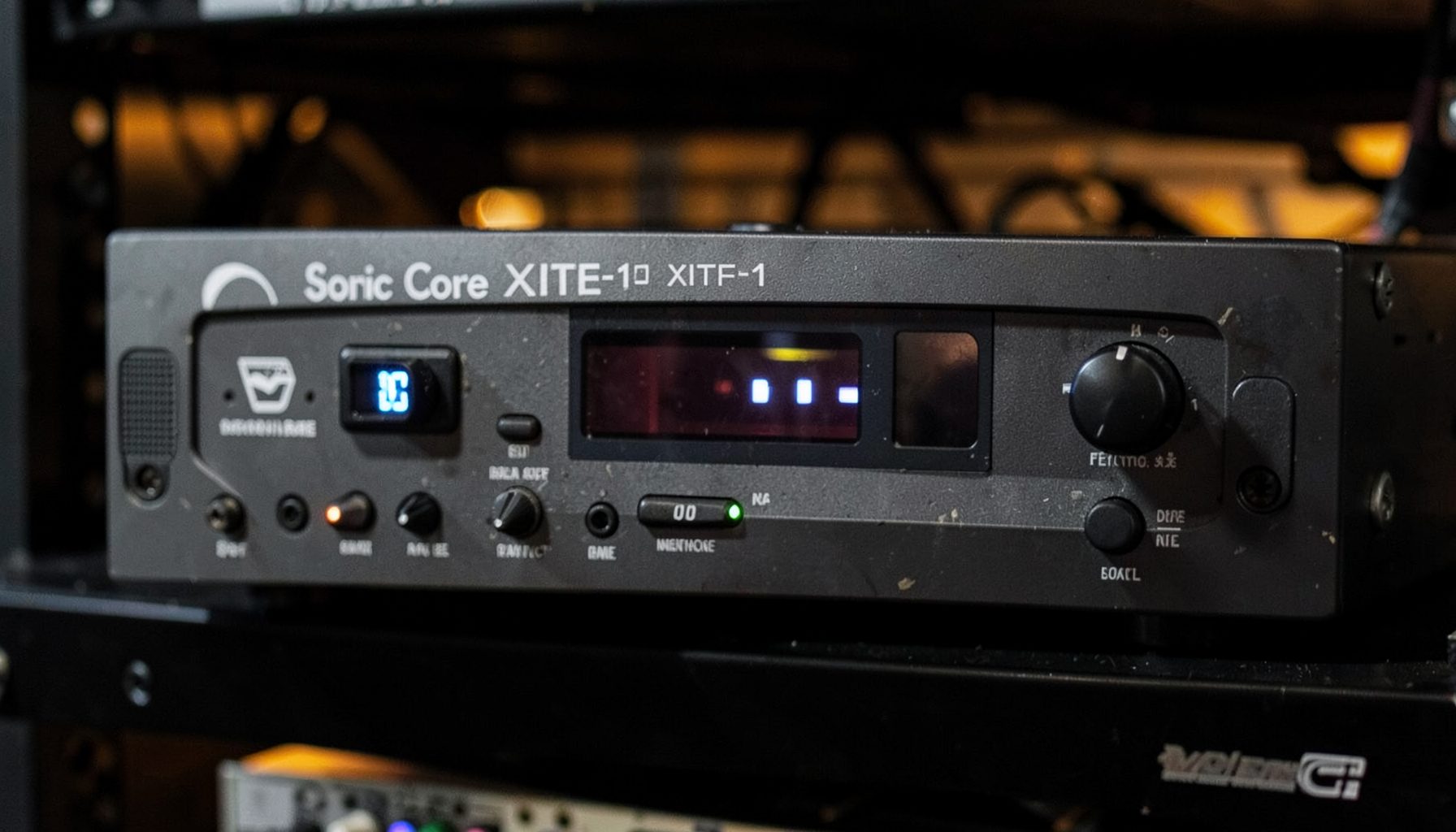 Close-up of a rack-mounted Sonic Core XITE-1 DSP unit with its front-panel display and knobs, set against a dark equipment rack with subtle amber glow from adjacent gear